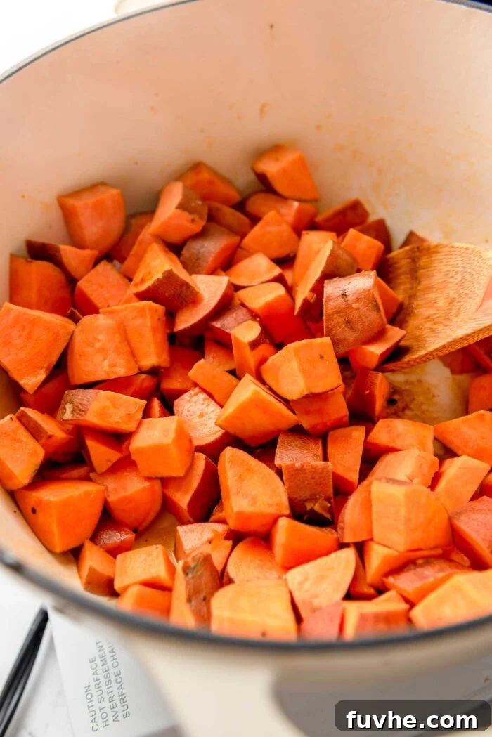 Cubes of sweet potato sizzling and browning in a large pot, with a wooden spoon for stirring, building delicious flavor for the curry.