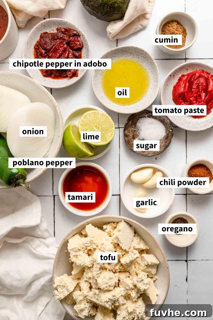 The ingredients for making copycat chipotle tofu sofritas gathered up in bowls on a counter. Each ingredient is labelled with text, showcasing extra-firm tofu, a poblano pepper, onion, chipotle in adobo, garlic cloves, tomato paste, tamari, spices, sugar, and lime.