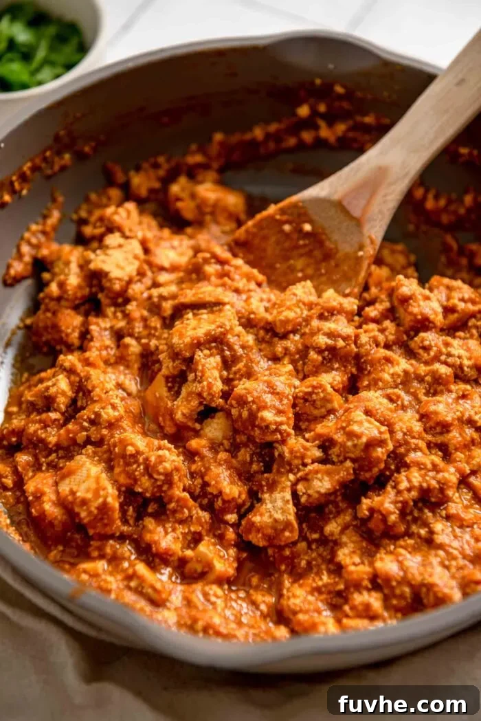 A wooden spoon mixing tofu sofritas, which is crumbled tofu in a spicy red sauce, in a skillet, demonstrating the simmering stage.