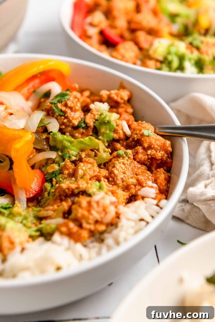 A close-up view of a Chipotle sofritas tofu burrito bowl, brimming with rice, cooked peppers and onions, creamy guacamole, and fresh cilantro. A fork rests in the bowl, with another bowl visible in the softened background.