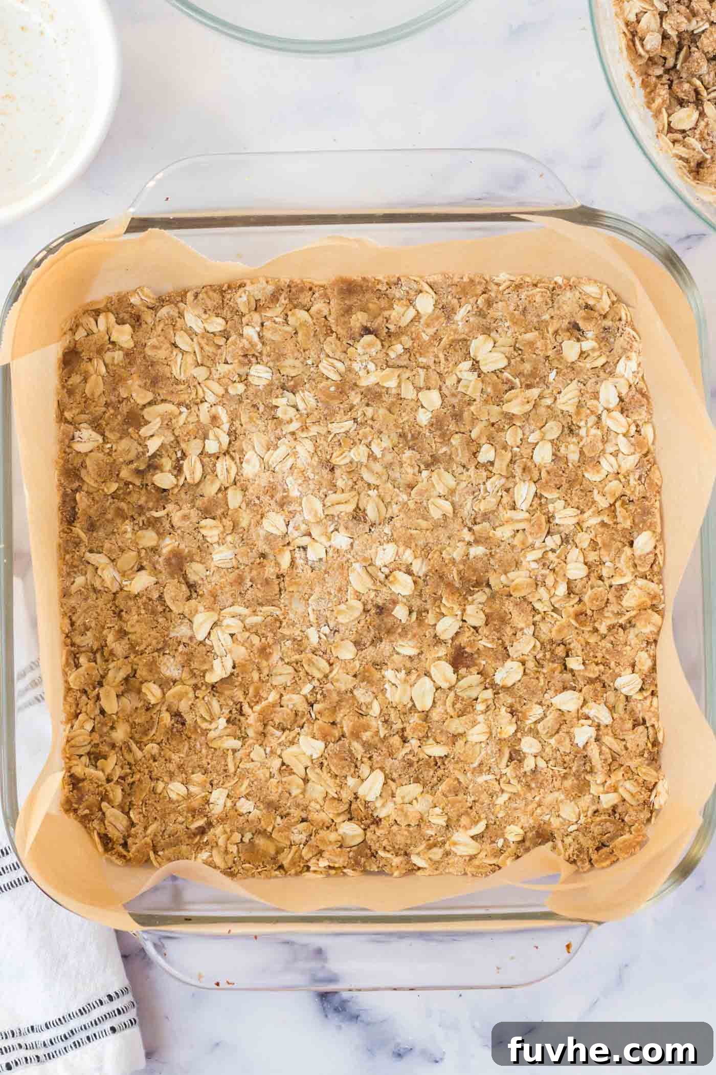 A thick oatmeal crust firmly pressed into the bottom of a square glass baking dish, meticulously lined with parchment paper.