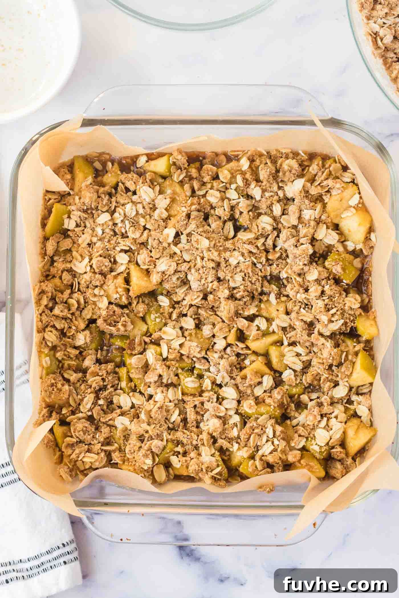 A generous layer of oatmeal streusel topping sprinkled artfully over the apple filling in a square glass baking dish, completing the bar assembly.