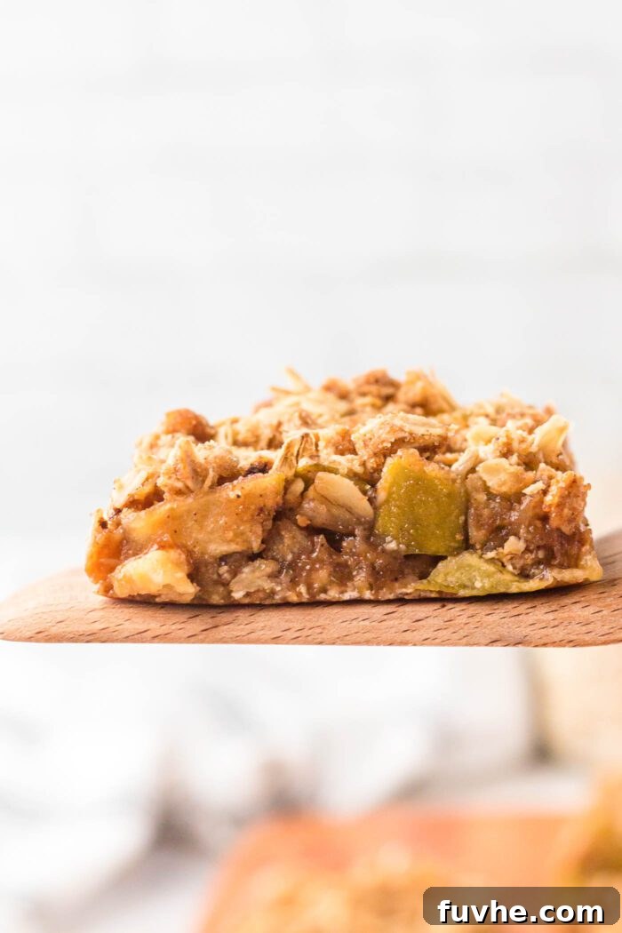 A single, perfectly baked apple pie crumble bar carefully held aloft on a wooden spatula, showcasing its layers.