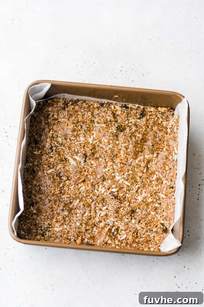 Paleo energy bars with coconut and hemp seeds pressed into a metal baking pan lined with parchment paper, ready for chilling.