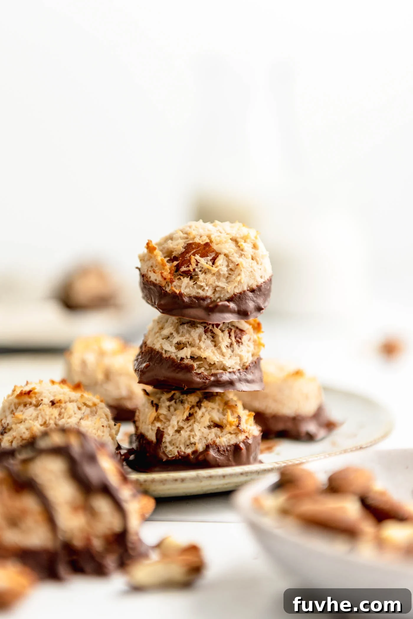 Plant-Powered Almond Coconut Dreams 2 Stack of 3 almond macaroons dipped in chocolate on a plate with more macaroons scattered around in the background and foreground.