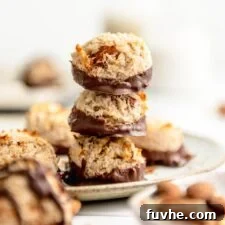 Stack of 3 coconut almond macaroons dipped in chocolate on a plate with more macaroons scattered around in the background and foreground.