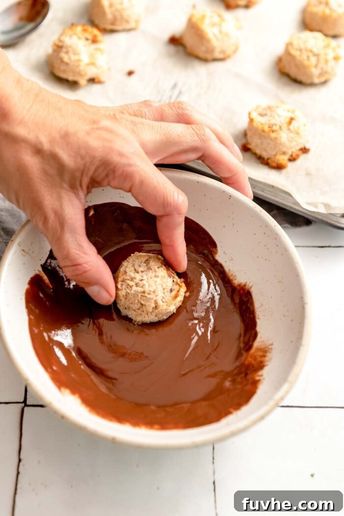 Plant-Powered Almond Coconut Dreams 8 Hand dipping an almond macaroon into a bowl of melted chocolate. There is a baking tray of macaroons in the background.