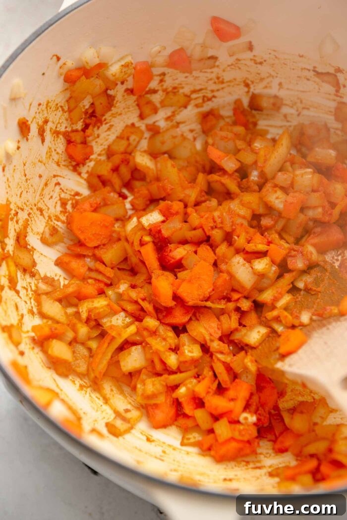 Chopped onion and carrot cooking in a spice mixture in a large soup pot with a wooden spoon.