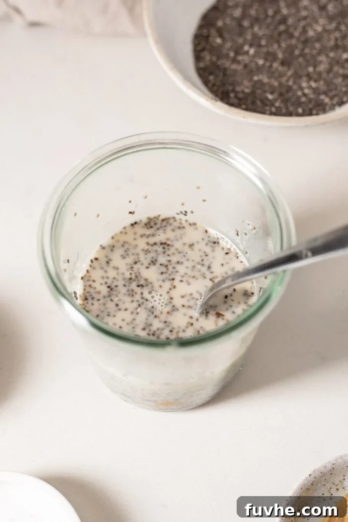 A close-up shot of chia seeds being mixed into plant-based milk in a glass jar with a spoon, showing the initial step of making chia pudding.
