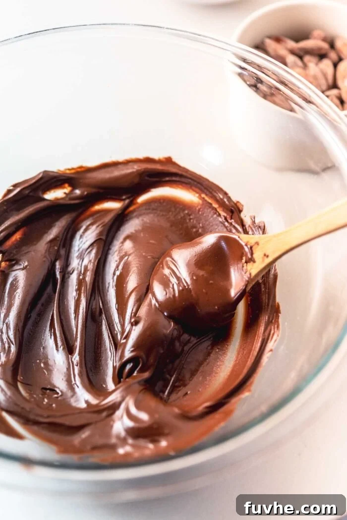 Irresistible Nutty Clusters 4 Smooth, glossy melted dark chocolate filling a clear glass mixing bowl, stirred lightly with a small wooden spoon, ready for the next ingredient.