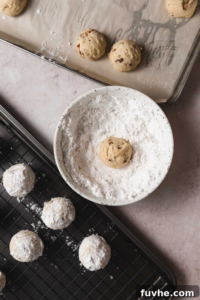 Melt-in-Your-Mouth Vegan Pecan Snowballs 8 A snowball cookie being coated in icing sugar in a small bowl. There is a sheet pan of cookies beside the bowl.