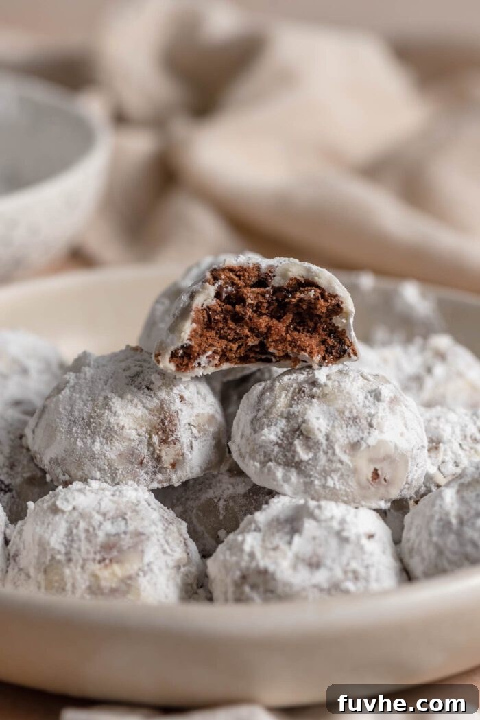 A bowl of icing sugar snowball cookies. The cookie on top has been bitten into so you can see the chocolate cookie with chocolate chips inside.