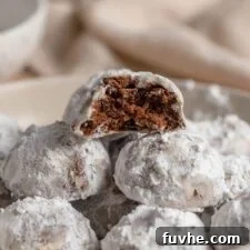 A bowl of icing sugar snowball cookies. The cookie on top has been bitten into so you can see the chocolate cookie with chocolate chips inside.