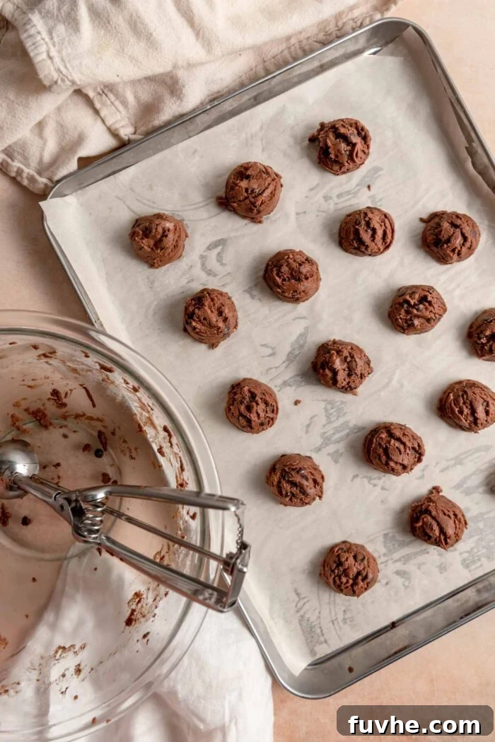 A cookie scoop resting in a glass mixing bowl beside a sheet pan of chocolate snowball cookies.