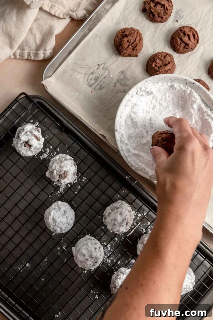 A hand dipping a chocolate snowball cookie in a bowl of icing sugar and placing them on a baking rack beside the bowl.