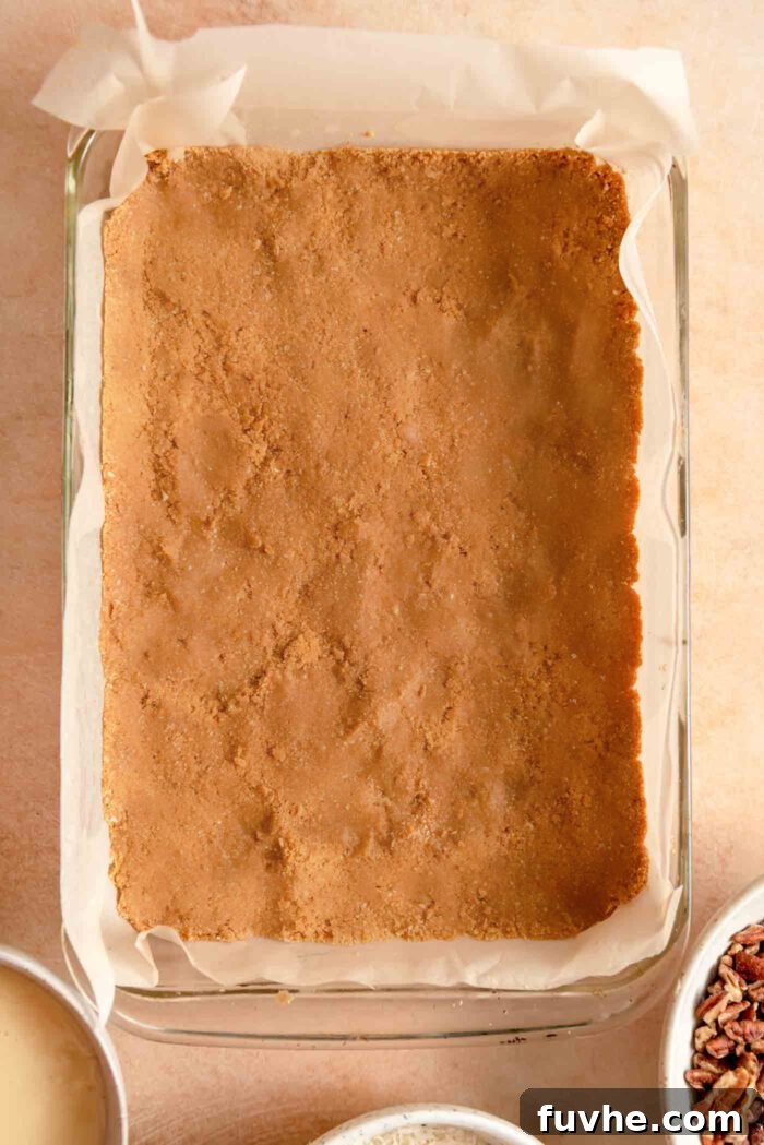 A smooth, even layer of graham cracker crust pressed firmly into a 9x13-inch baking pan, lined with parchment paper, ready for the next layers.