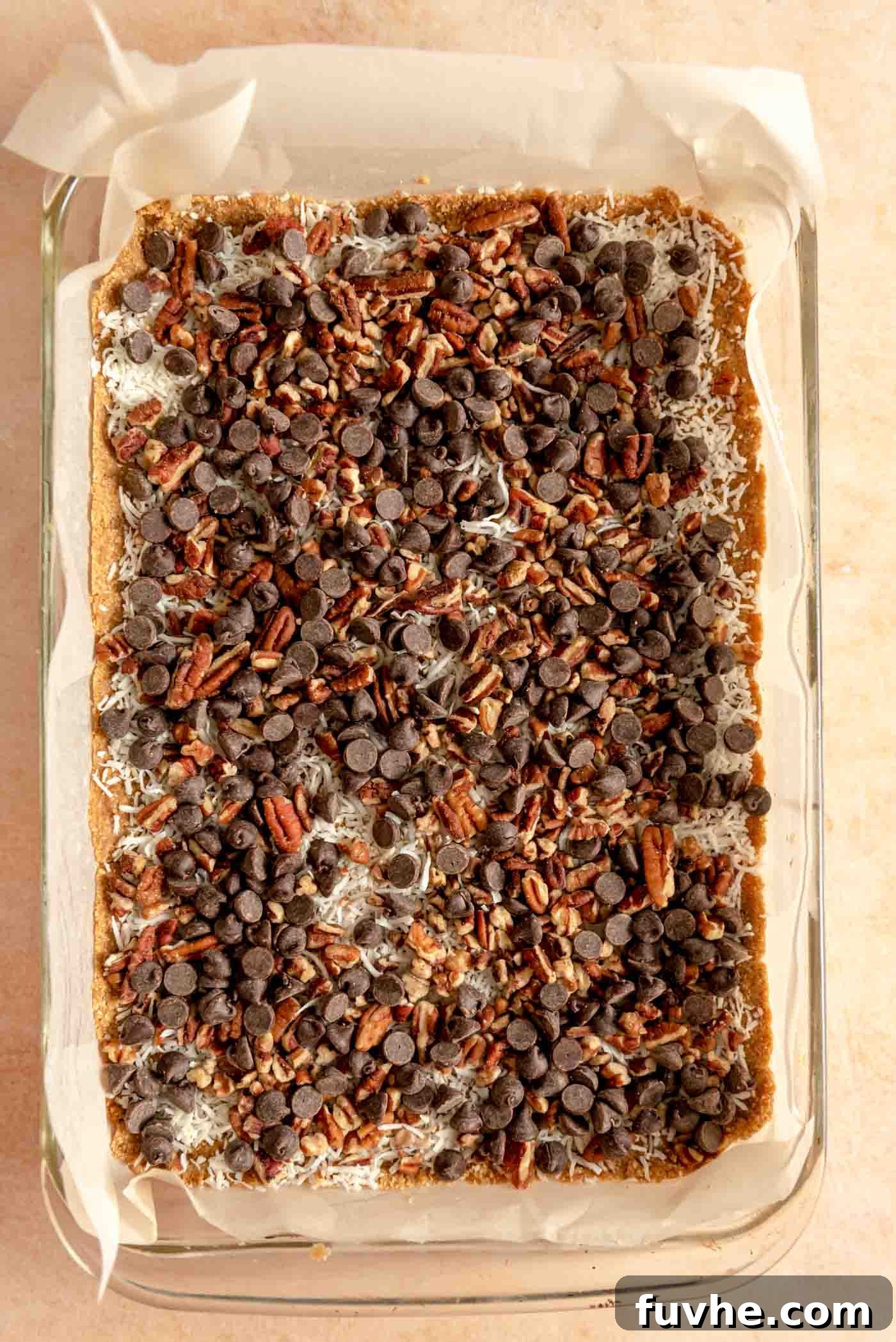 Dairy-free chocolate chips, chopped pecans, and shredded coconut are generously sprinkled over the graham cracker crust in a glass baking dish, forming the first set of layers for the magic bars.
