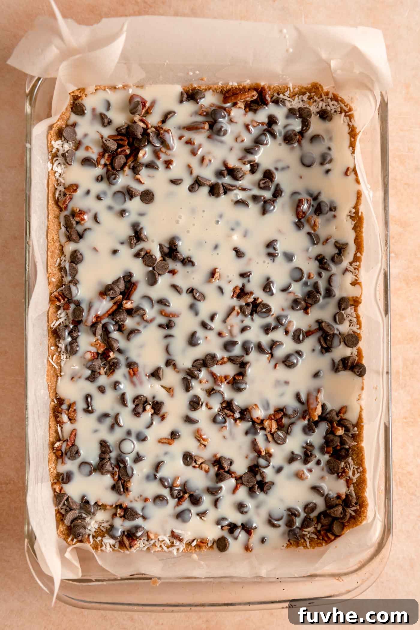 Sweetened coconut condensed milk being poured over the layers of chocolate chips, pecans, and coconut in a 9x13-inch glass baking pan, ready for baking.