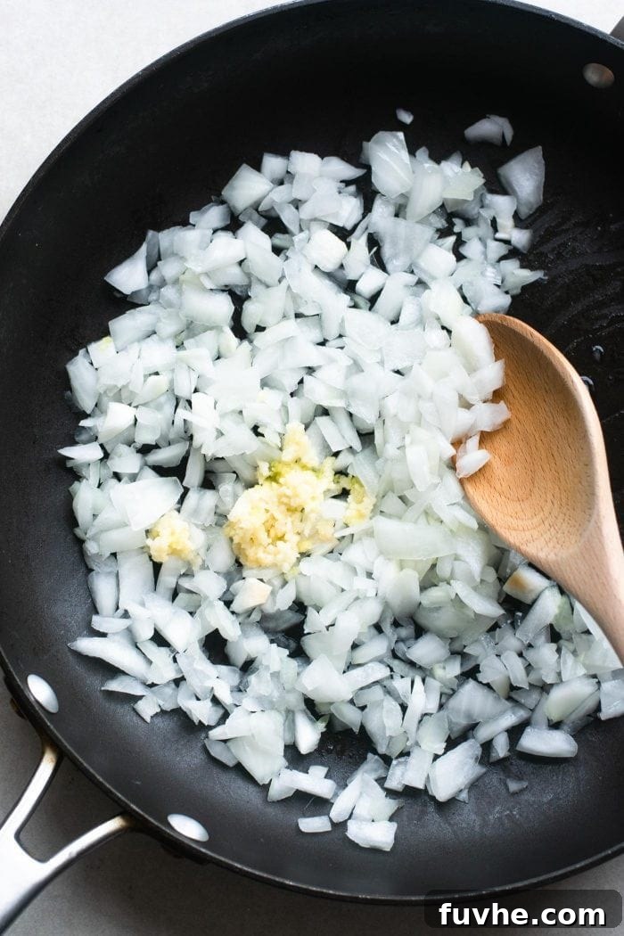 Finely chopped onions and minced garlic sizzling in a skillet with a wooden spoon, releasing their fragrant aromas as they soften for the cauliflower rice dish.