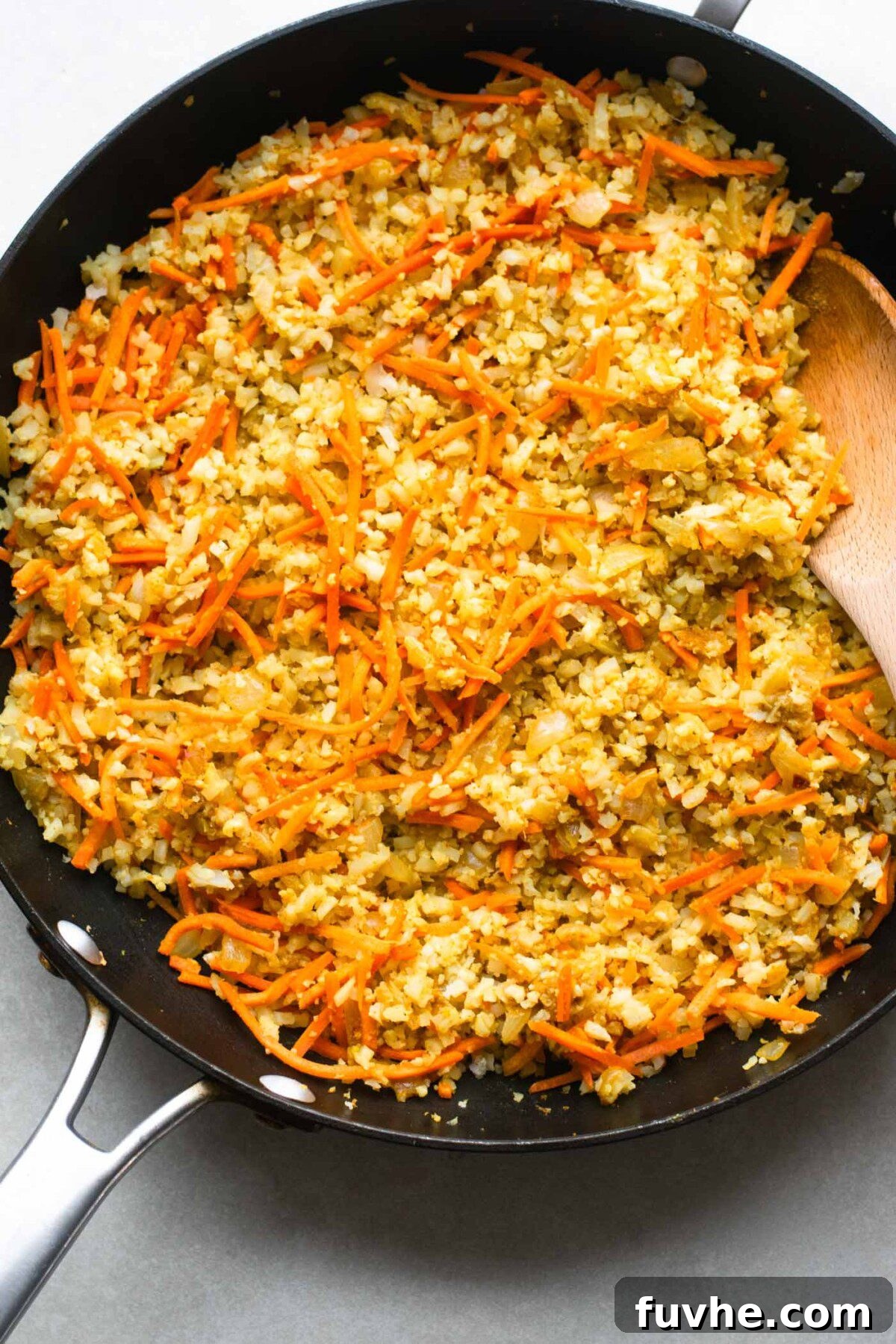 A large non-stick pan filled with riced cauliflower and grated carrots stir-frying with aromatic spices, onions, and garlic, creating a colorful and healthy base for the dish.