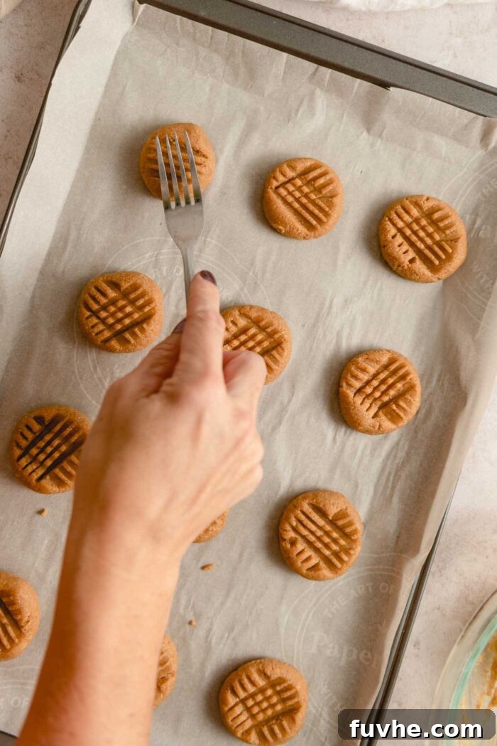 A hand using a fork to press a crisscross pattern onto peanut butter cookies on a baking tray.