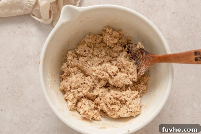 A bowl filled with vegan coconut oatmeal cookie dough, featuring visible oats and coconut flakes, being mixed into a uniform consistency with a wooden spoon.