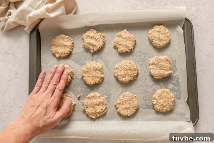 A hand gently flattening a raw coconut oatmeal cookie on a baking sheet, which is already populated with 12 other perfectly portioned cookie dough balls, ready for baking.