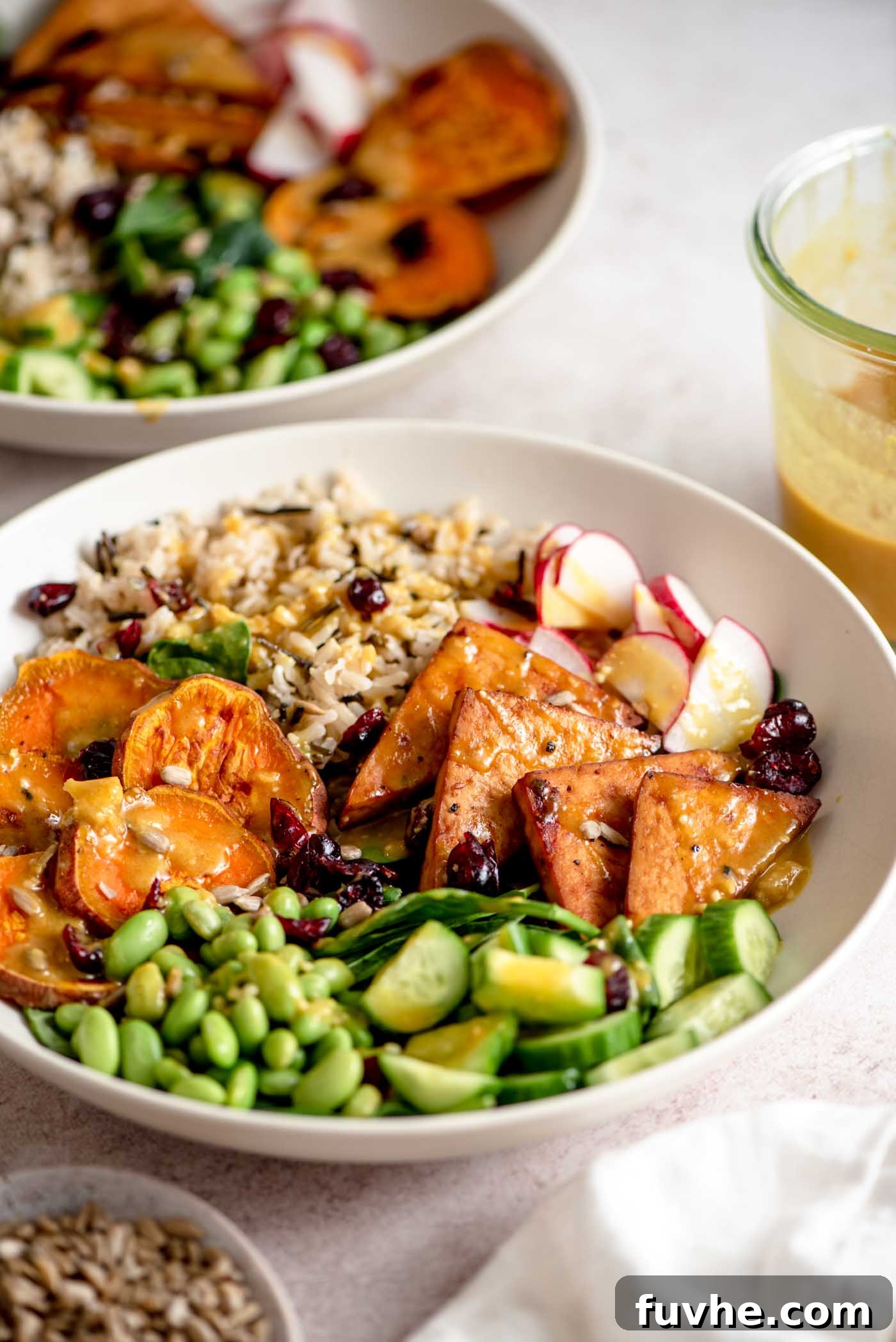 A colourful veggie power bowl with sweet potato, tofu, rice, edamame, cucumber, radish and spinach all topped with sauce. There's another bowl in the background.
