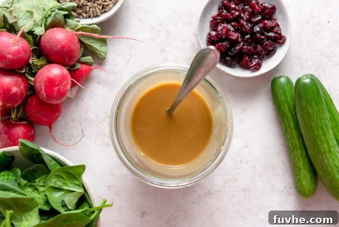 Creamy miso tahini dressing in a small glass container with a spoon in it surrounded by some spinach in a bowl, radishes, cucumber and dried cranberries in a small dish.
