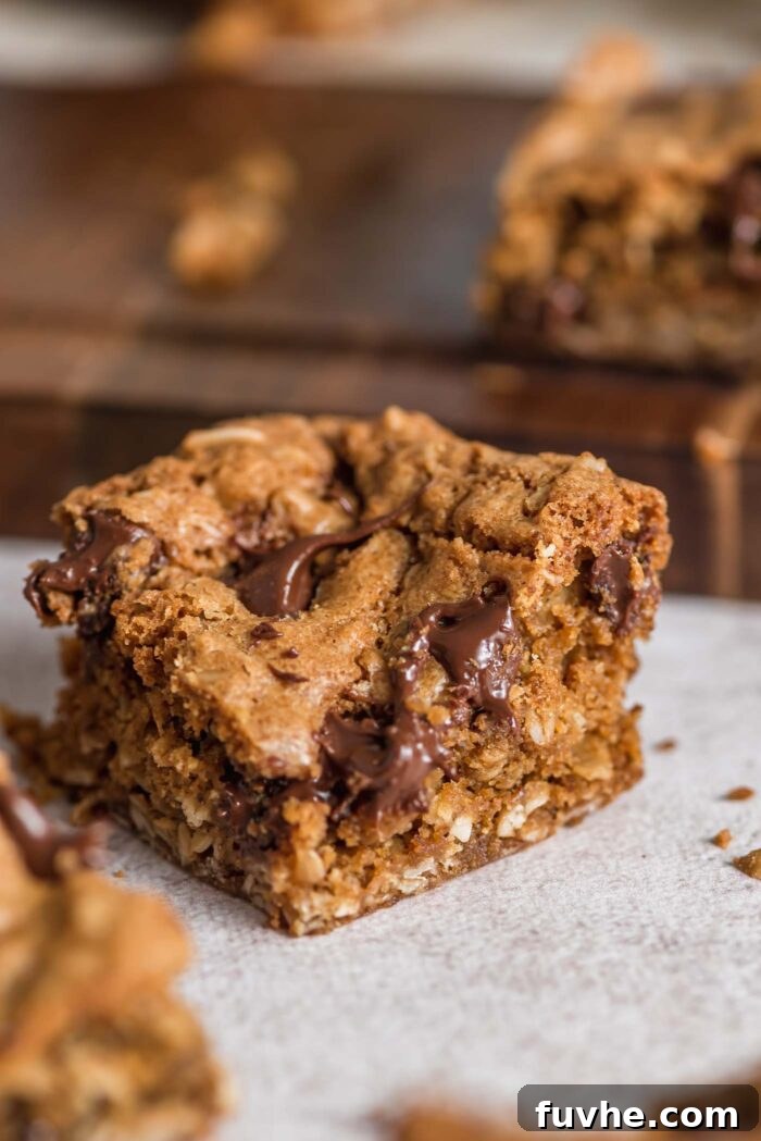 Wholesome Vegan Oatmeal Bars 10 An oatmeal cookie bar sitting on a counter with a cutting board behind it with more bars on it. You can see the texture and melted chocolate chips inside the bar.