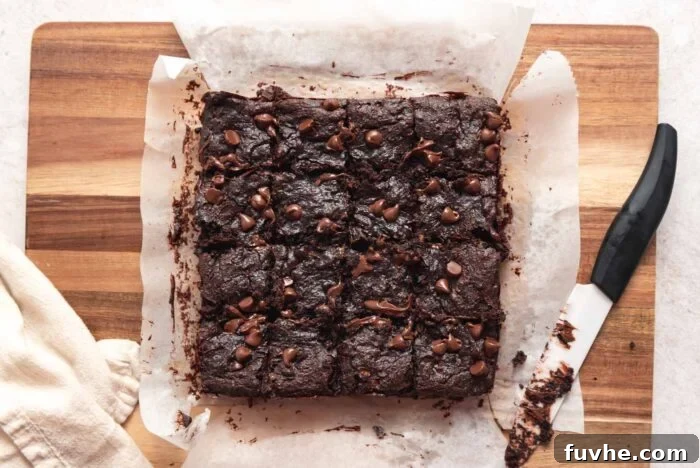 A batch of chocolate chip brownies cut into 16 squares on a wooden cutting board with a knife resting on the edge of it.