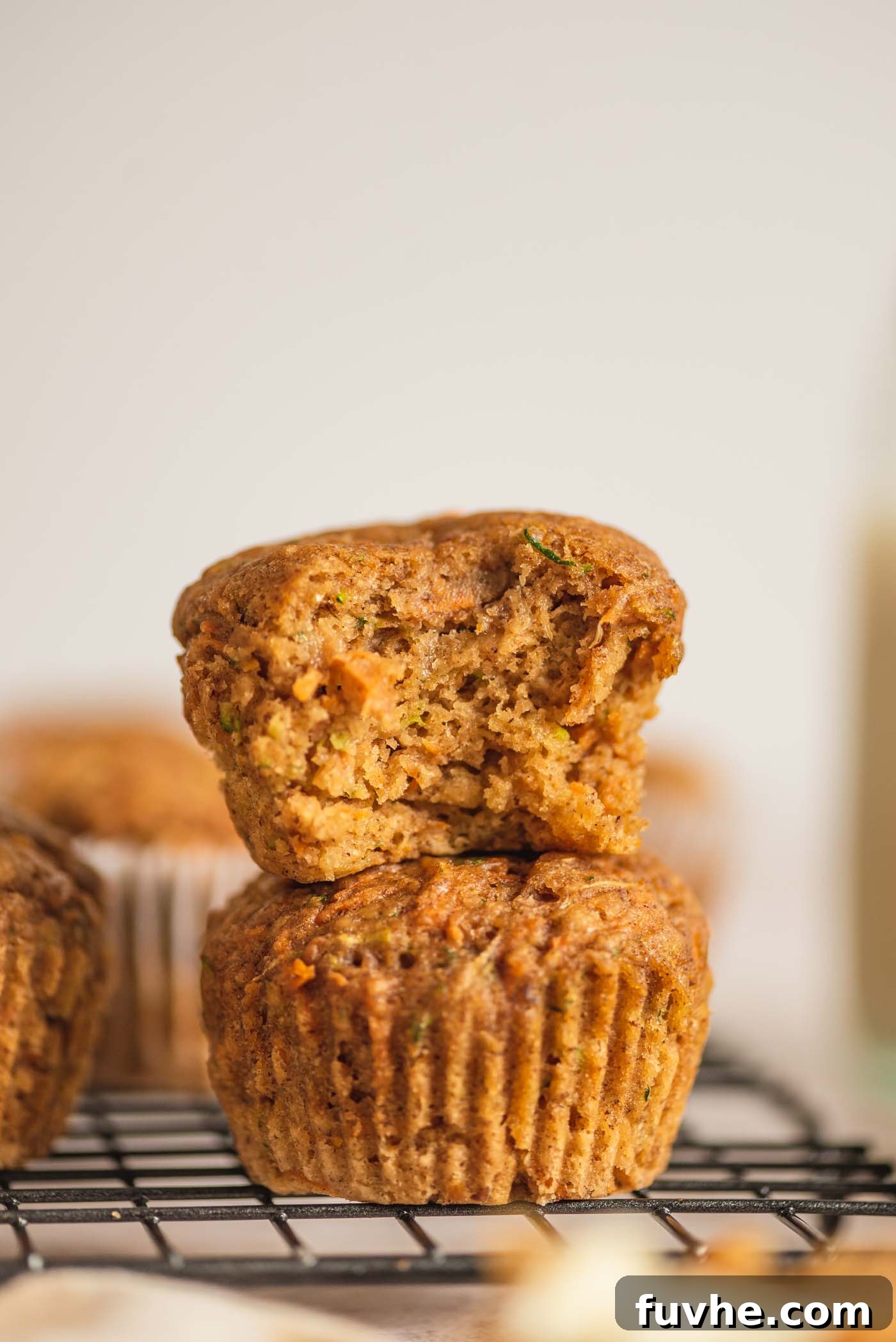 Two healthy vegan carrot zucchini muffins stacked. The top muffin has a bite taken out, revealing its moist, tender crumb and visible flecks of grated carrot and zucchini.