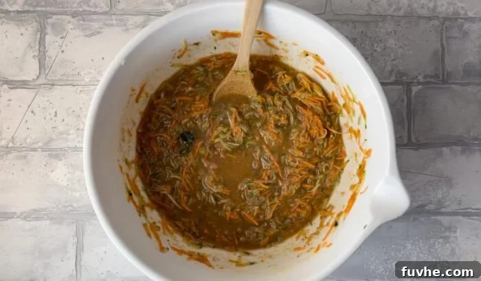 Raw carrot zucchini muffin batter in a mixing bowl with a wooden spoon, showcasing the rich texture before baking.