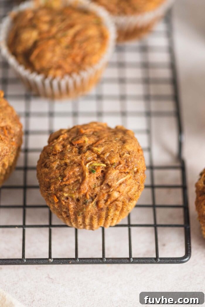 A small, perfectly baked carrot zucchini muffin resting on the edge of a baking rack, with several other muffins cooling in the background.