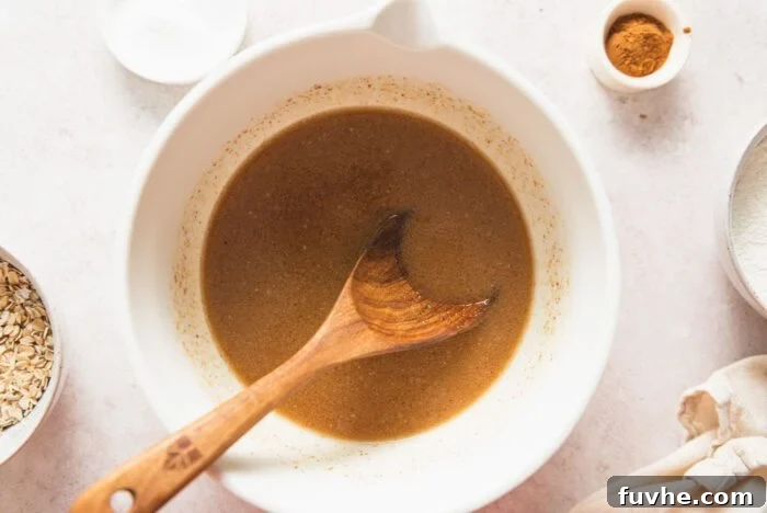 Maple syrup, brown sugar, applesauce and almond milk mixed into a brown mixture in a mixing bowl with a wooden spoon resting in it.