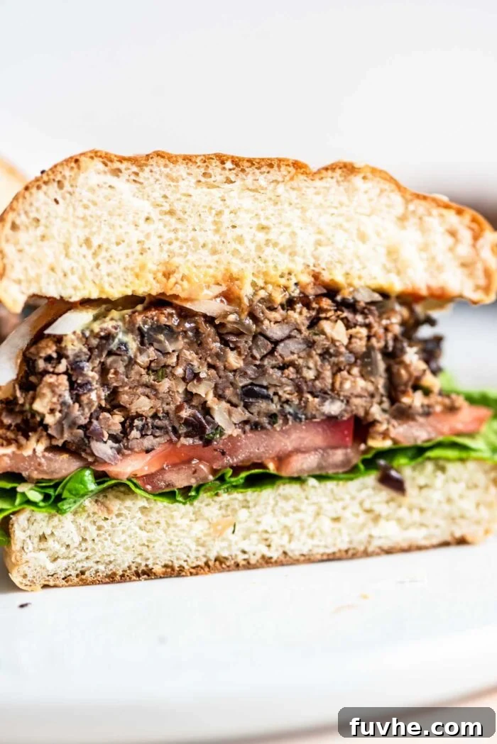 A perfectly assembled vegan mushroom burger on a toasted bun, garnished with fresh tomato, crisp lettuce, red onion, and a swirl of ketchup and mustard. The burger is cut in half to showcase its hearty layers.