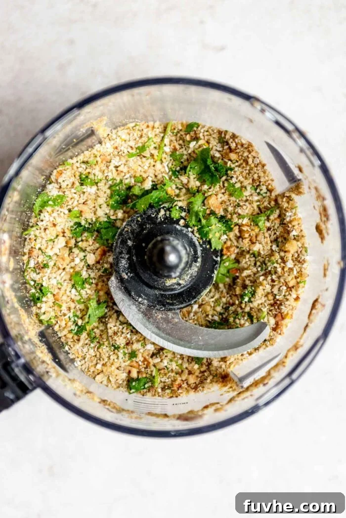 A close-up view of the food processor bowl containing a blended mixture of oats, walnuts, panko breadcrumbs, ground flax, nutritional yeast, and fresh cilantro or parsley, partially pulsed.