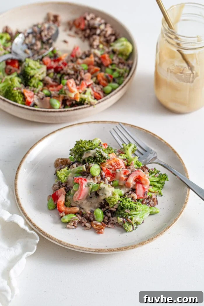 Broccoli, bell pepper, raisin, rice and edamame salad on a plate with a spoon. Bowl of salad in background.