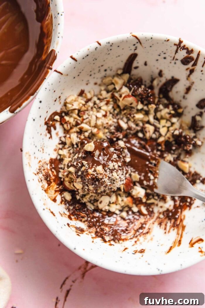 Rolling a chocolate-coated truffle in chopped hazelnuts in a small bowl.