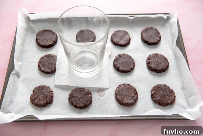 A glass sitting on a piece of parchment paper on one small, round cookie of a tray of 12 cookies. The glass is being used to flatten the cookies to an even thickness for baking.