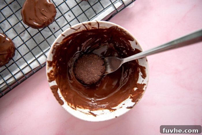 A small chocolate cookie being carefully dipped into a bowl of melted mint chocolate coating using a fork.