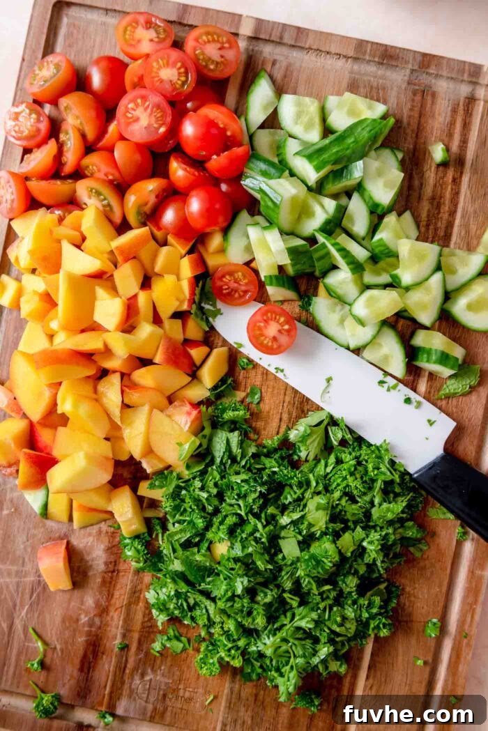 Chopped fresh herbs, nectarine, cherry tomato and cucumber on a cutting board with a knife.