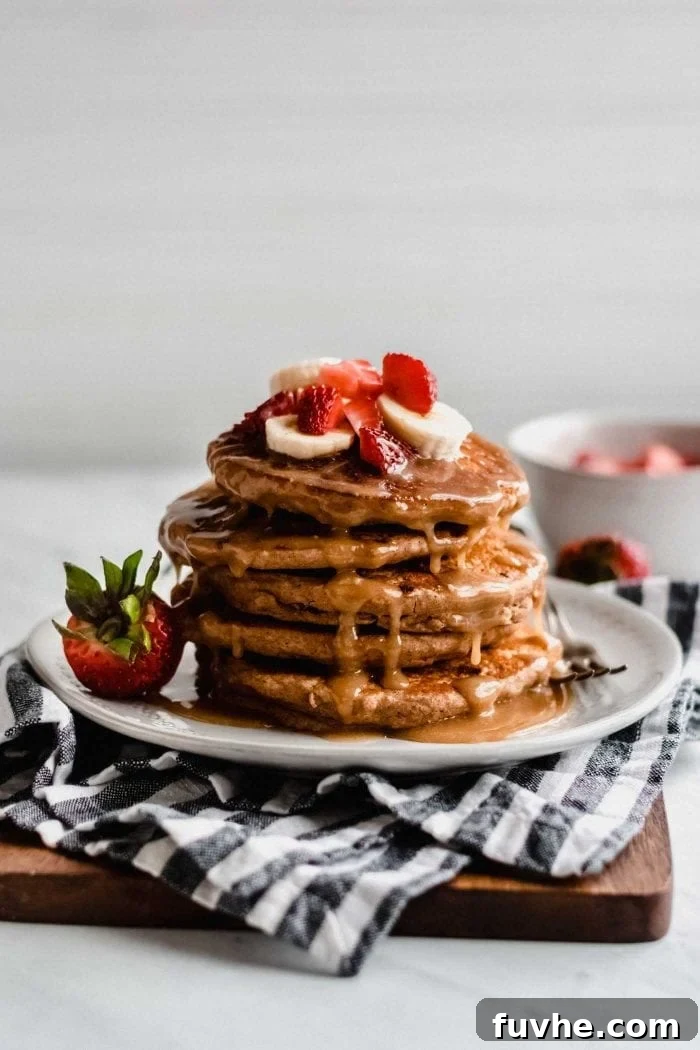 A stack of golden-brown vegan peanut butter banana pancakes, generously topped with vibrant sliced bananas, fresh red strawberries, and a luscious drizzle of homemade peanut butter sauce. The plate is garnished with extra strawberries, inviting a closer look.