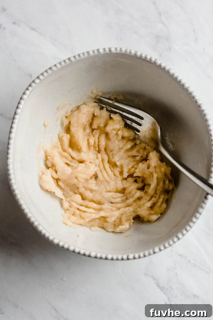 A bowl containing perfectly mashed banana, ready for the next steps of the pancake batter preparation, with a fork resting beside it.