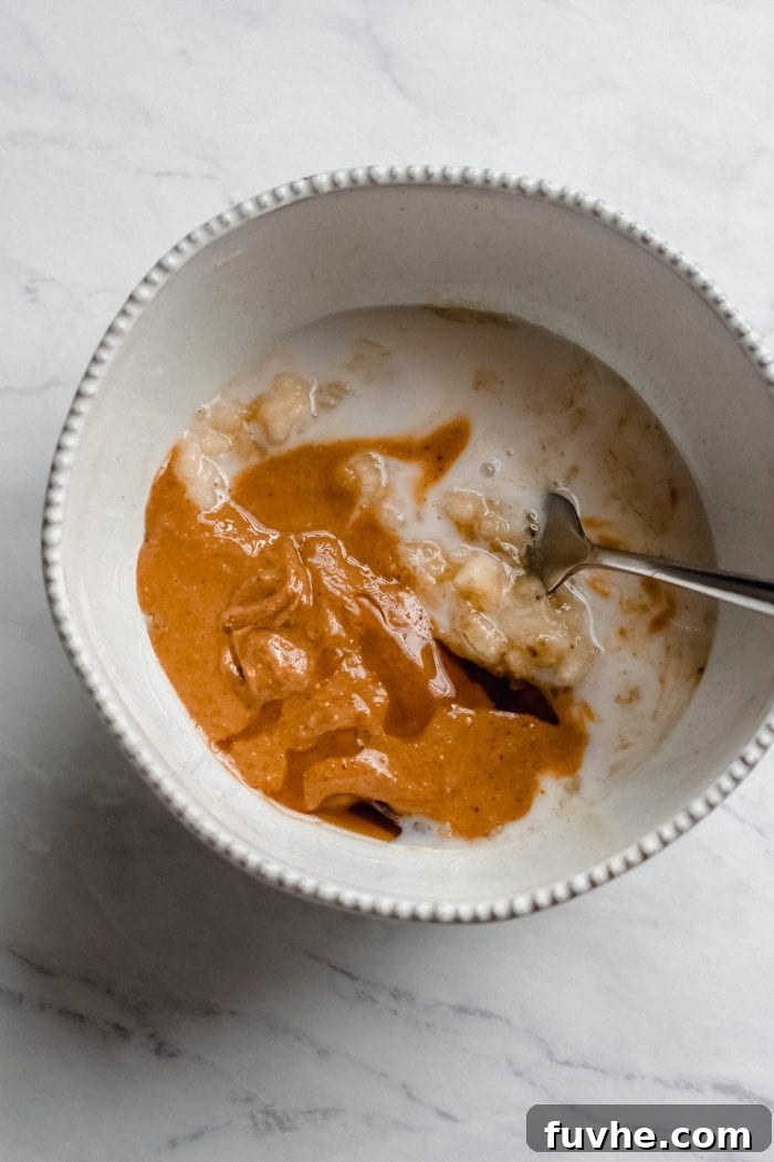 A mixing bowl filled with creamy almond milk, mashed banana, and smooth peanut butter, being expertly blended together with a fork, creating a homogenous wet mixture for the pancake batter.