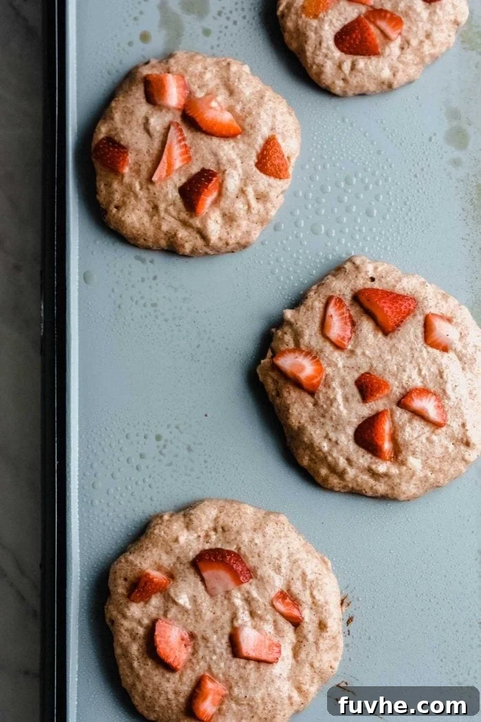 Several vegan peanut butter banana pancakes dotted with strawberry pieces sizzling gently on a hot pancake griddle, showcasing the cooking process before flipping.