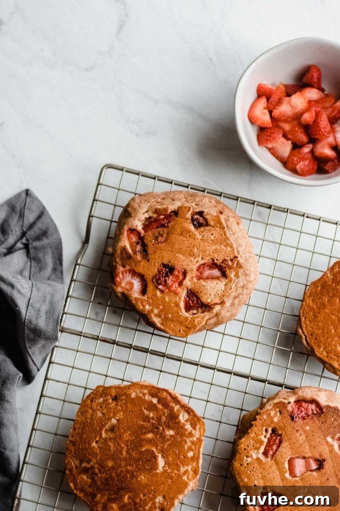 A cooling rack neatly displaying freshly cooked vegan peanut butter banana pancakes, each infused with strawberries. Beside the rack sits a small bowl of vibrant chopped strawberries, ready for topping.