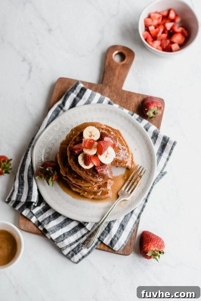 An inviting overhead shot showcasing a meticulously arranged stack of strawberry peanut butter banana pancakes, artfully placed on a plate over a rustic cutting board. The pancakes are generously adorned with fresh, vibrant berries, perfectly sliced bananas, and a glistening pour of maple syrup, capturing a moment of pure breakfast bliss.