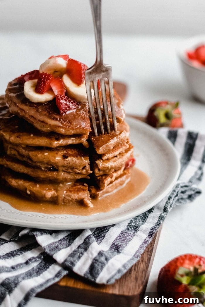 A close-up shot of a fork expertly piercing a tall stack of fluffy vegan peanut butter banana pancakes, generously adorned with fresh strawberries and a delectable peanut butter sauce, inviting a delicious bite.