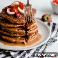 A close-up shot of a fork expertly piercing a tall stack of fluffy vegan peanut butter banana pancakes, generously adorned with fresh strawberries and a delectable peanut butter sauce, inviting a delicious bite.